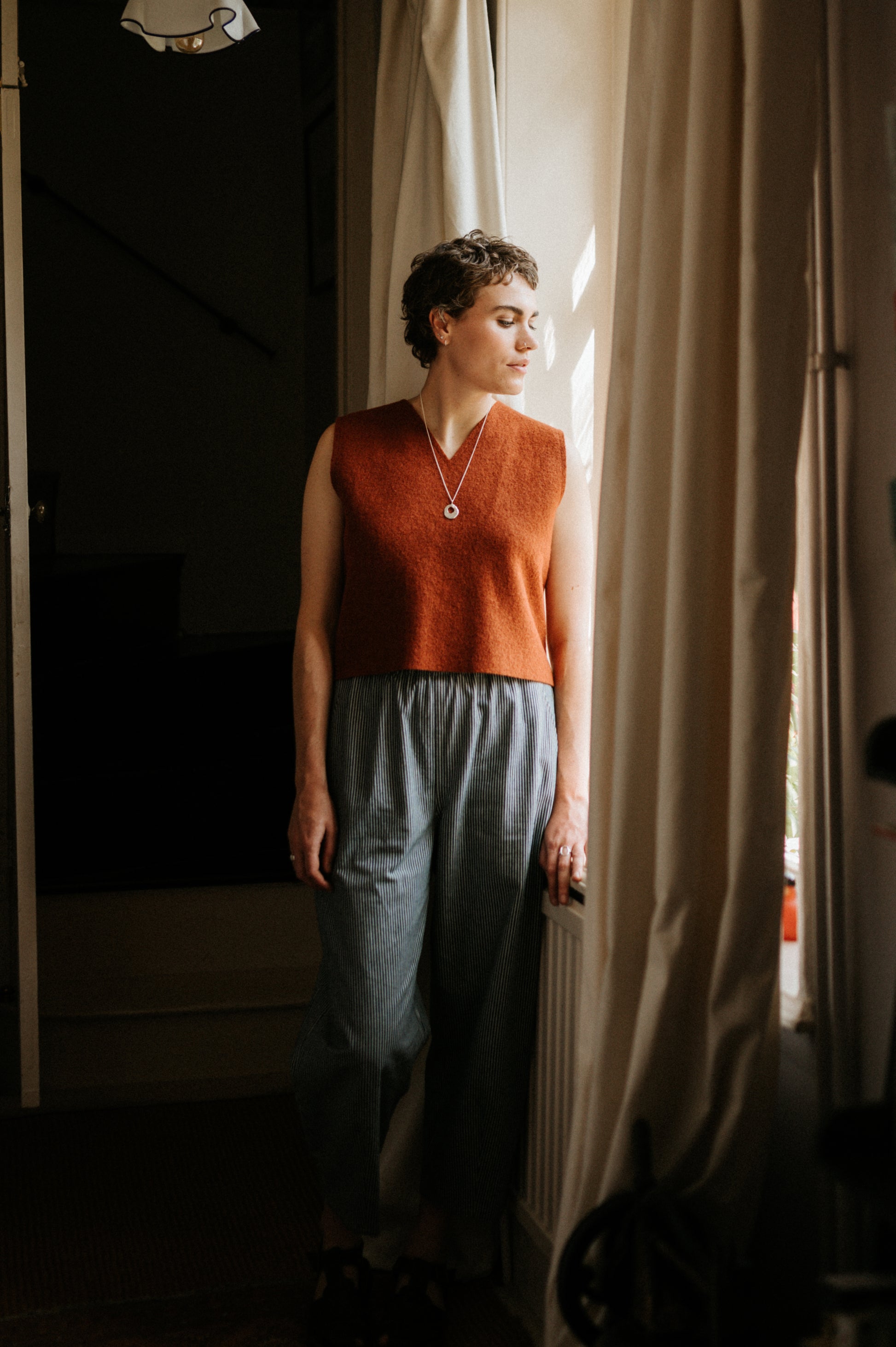 Person wearing an orange sleeveless boiled wool vest and blue trousers standing in a doorway.