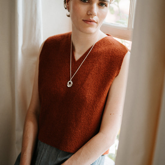 Woman wearing a rust-colored sleeveless vest sitting by a window.