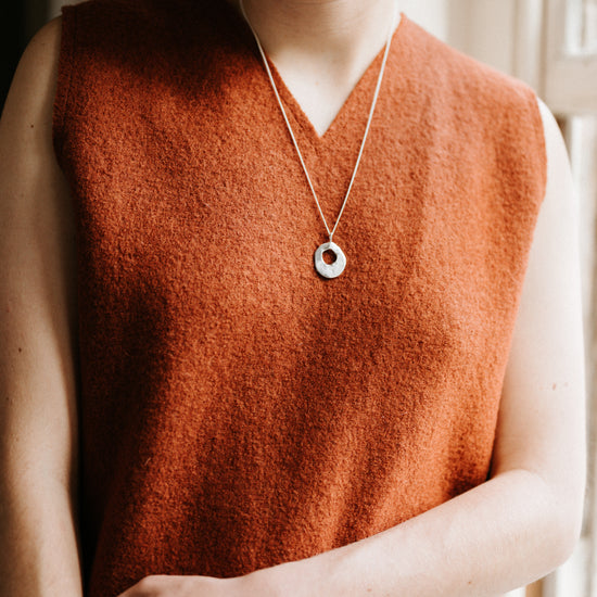 Person wearing a rust-colored sleeveless vest with a necklace, standing indoors.