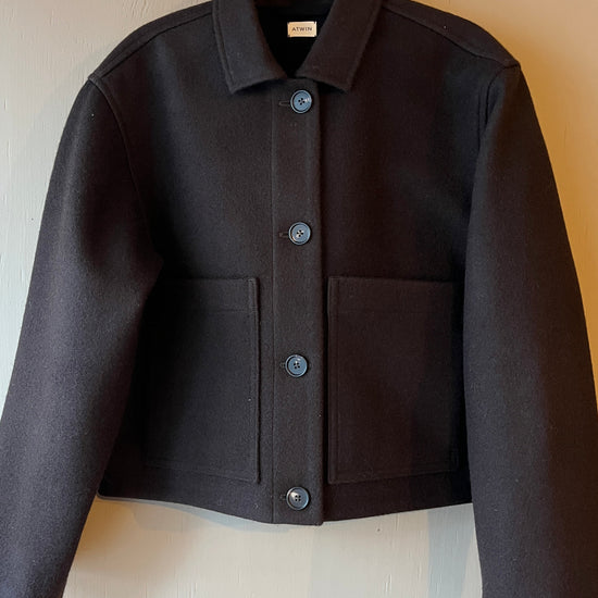 Brown wool jacket with brown buttons, long sleeves, collar and front patch pockets on a light grey surface, hanging from a wooden peg hook.