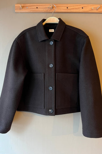 Brown wool jacket with brown buttons, long sleeves, collar and front patch pockets on a light grey surface, hanging from a wooden peg hook.