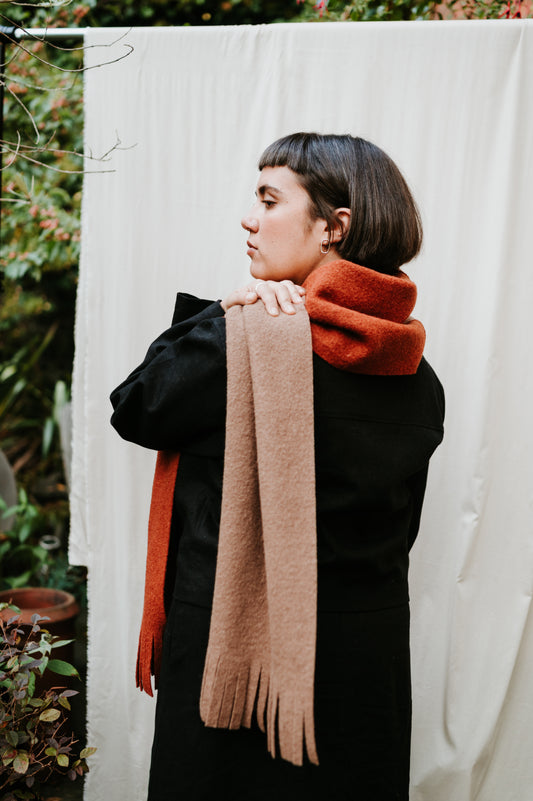 Person wearing a black coat with a brown scarf and orange sweater against a white curtain.