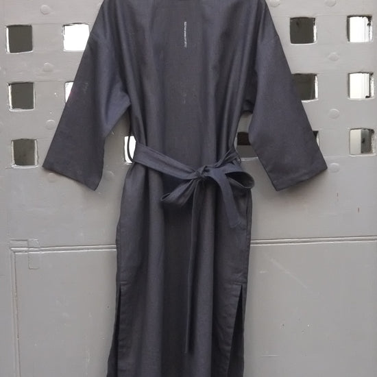 A black long-sleeve tunic dress with a matching waist tie belt, hanging on a wooden hanger against a gray metal door with a grid pattern.