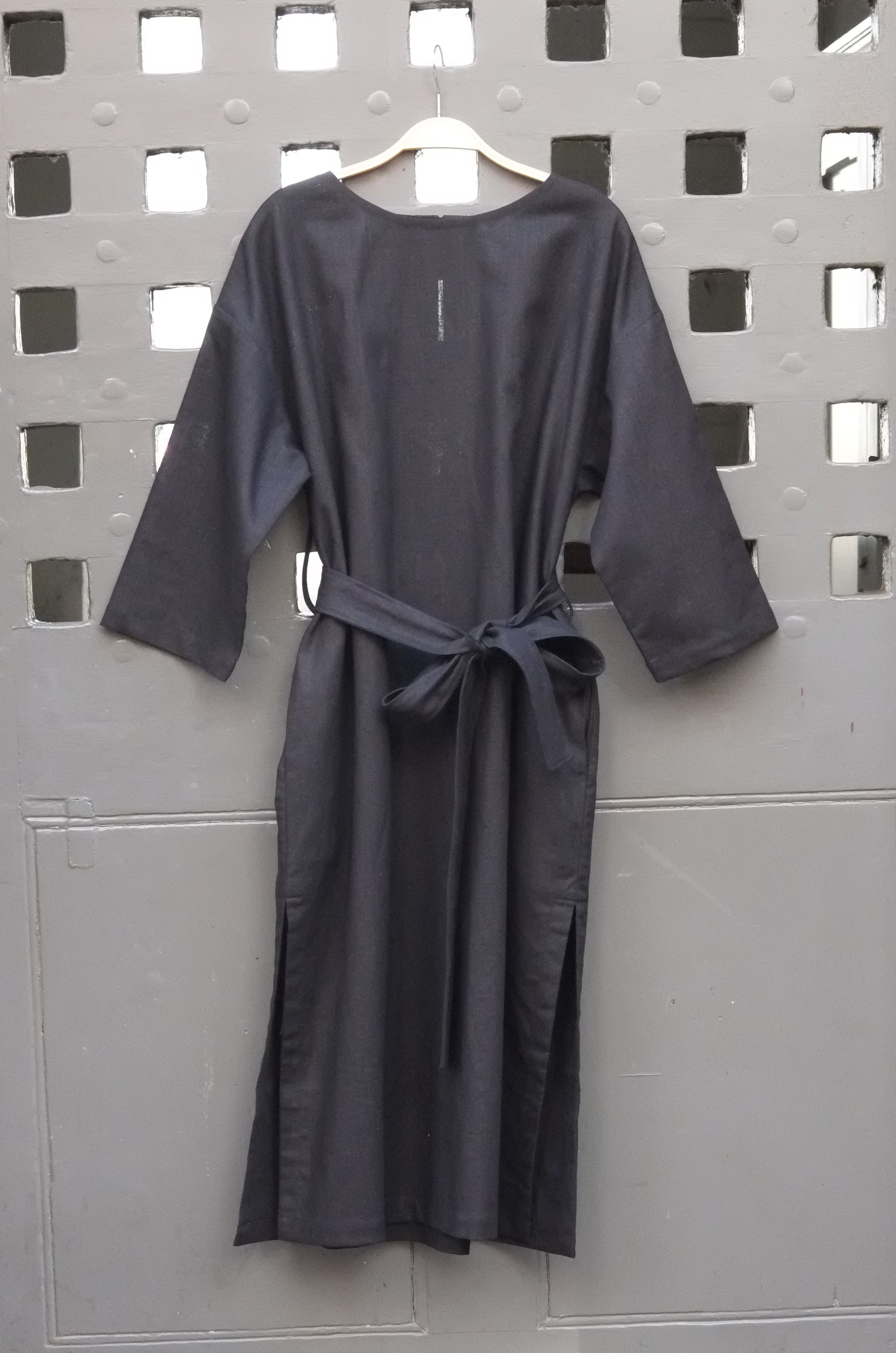 A black long-sleeve tunic dress with a matching waist tie belt, hanging on a wooden hanger against a gray metal door with a grid pattern.