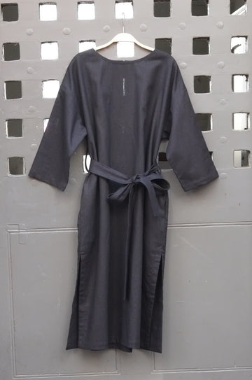 A black long-sleeve tunic dress with a matching waist tie belt, hanging on a wooden hanger against a gray metal door with a grid pattern.