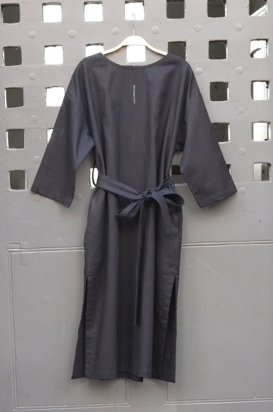 A black long-sleeve tunic dress with a matching waist tie belt, hanging on a wooden hanger against a gray metal door with a grid pattern.