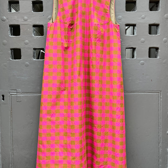 A sleeveless pink and  green checkered midi dress hanging on a wooden hanger against a grey door.