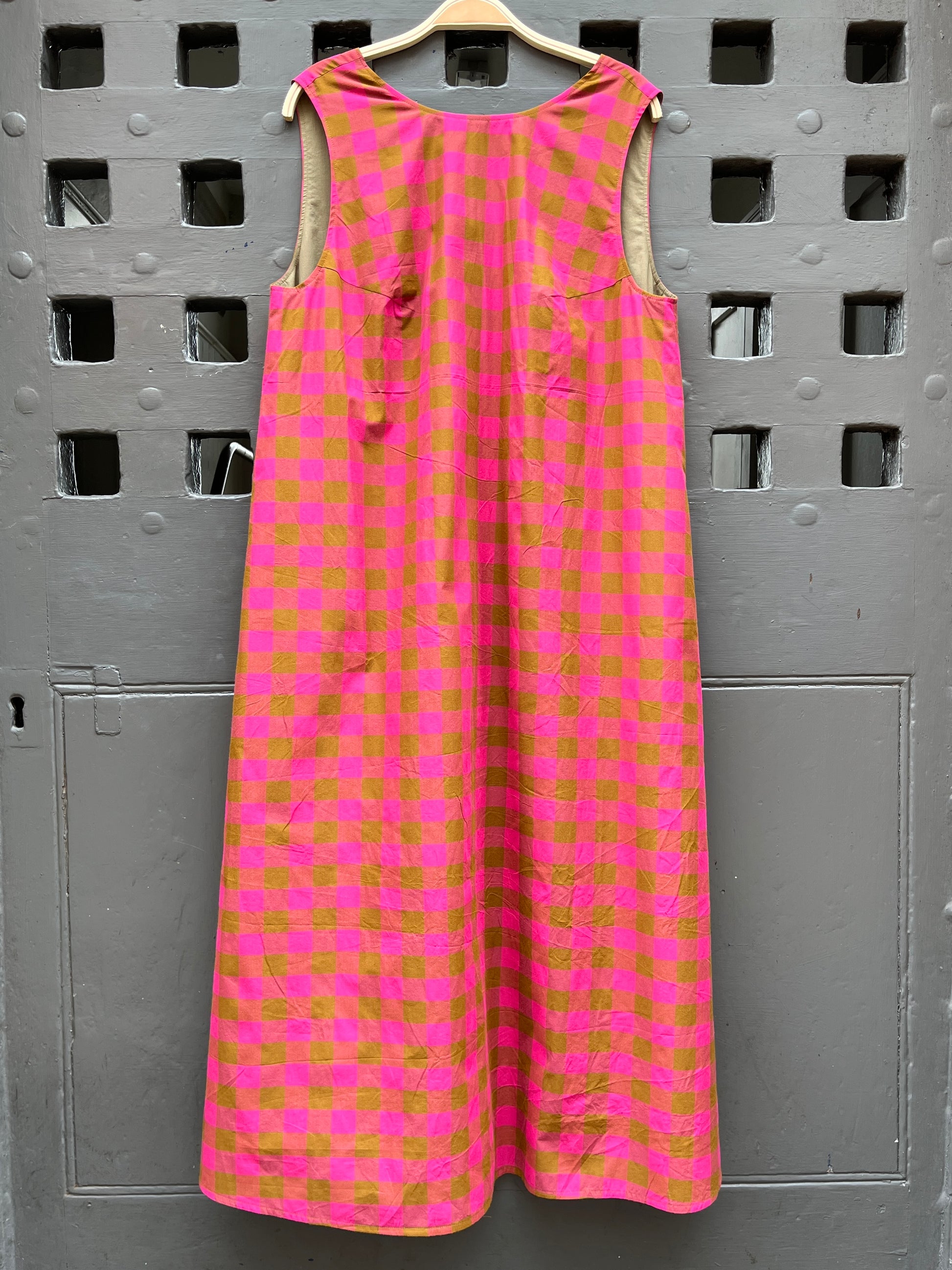 A sleeveless pink and  green checkered midi dress hanging on a wooden hanger against a grey door.