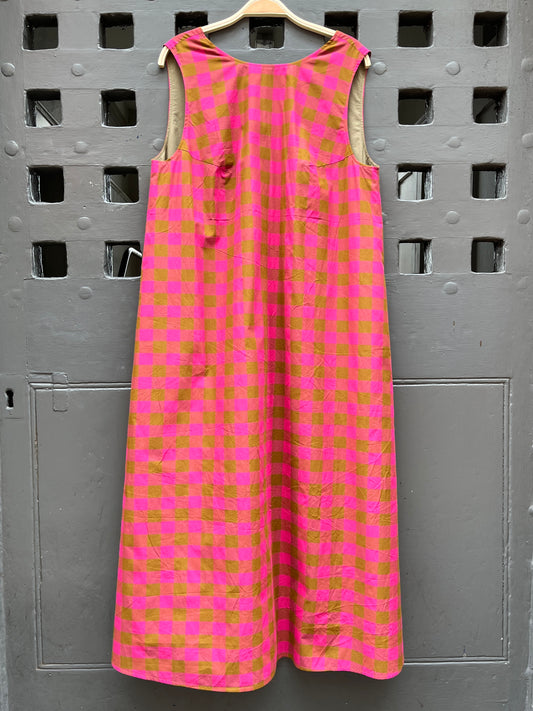 A sleeveless pink and  green checkered midi dress hanging on a wooden hanger against a grey door.