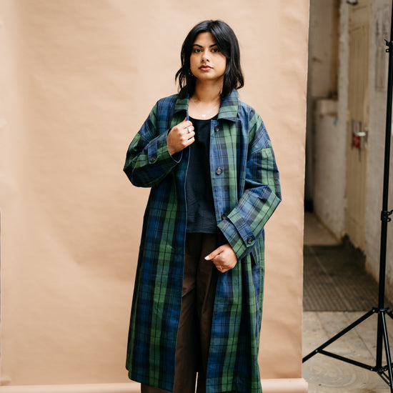 A woman stands in a studio setting wearing a long green and blue plaid coat over a dark top and brown wide-leg pants, with red shoes. A beige paper backdrop is behind her, with a lighting stand visible on the right.