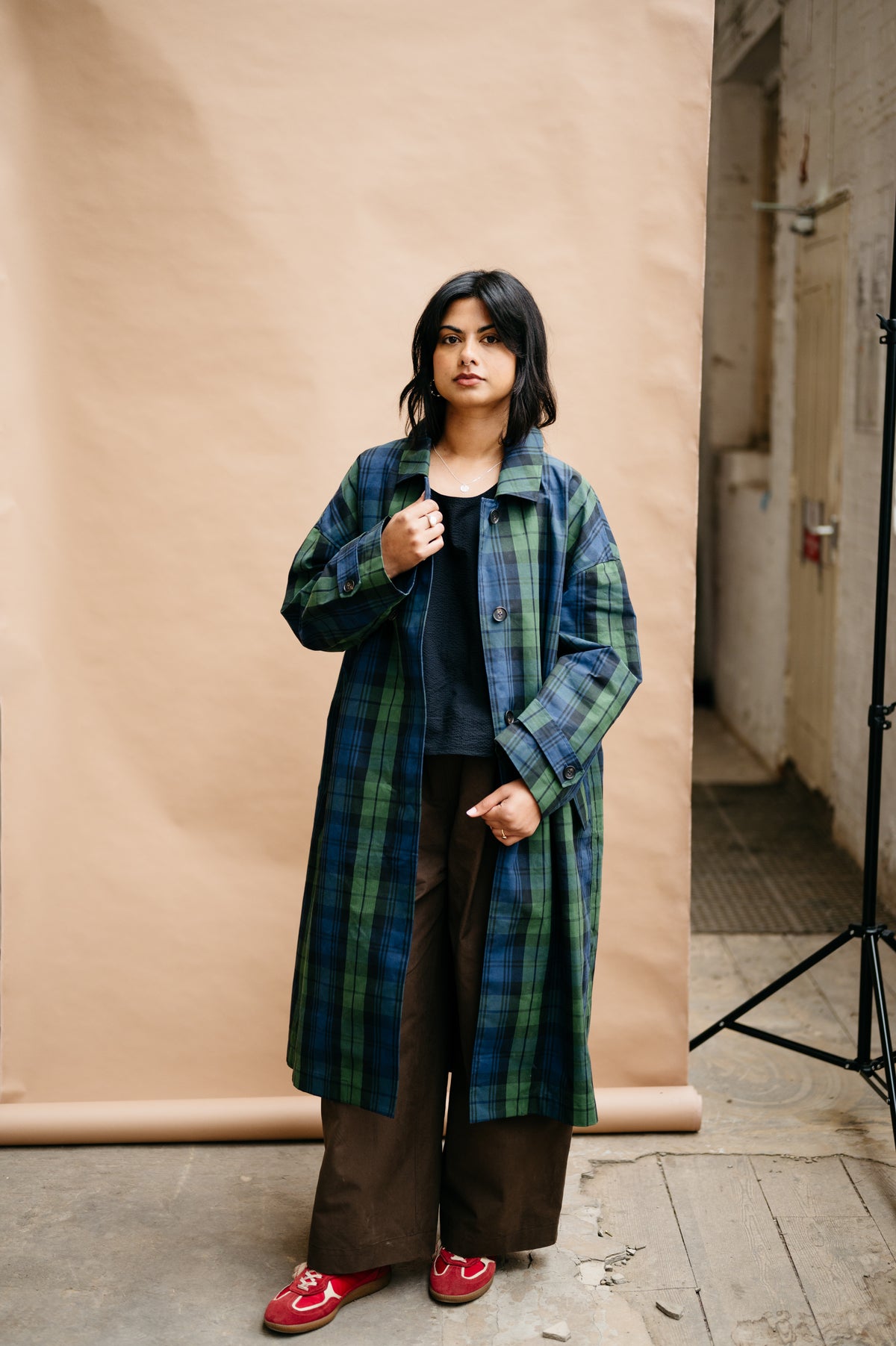 A woman stands in a studio setting wearing a long green and blue plaid coat over a dark top and brown wide-leg pants, with red shoes. A beige paper backdrop is behind her, with a lighting stand visible on the right.