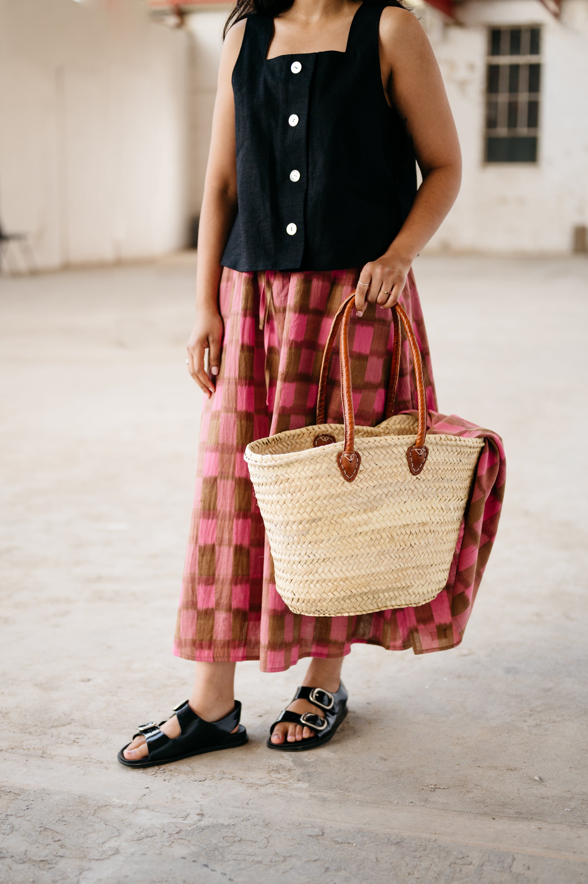 Person wearing a black top and pink plaid skirt holding a straw bag.