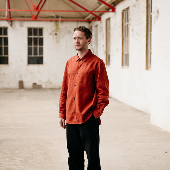 Man wearing an orange shirt and black pants standing in a large, empty warehouse with red metal beams.
