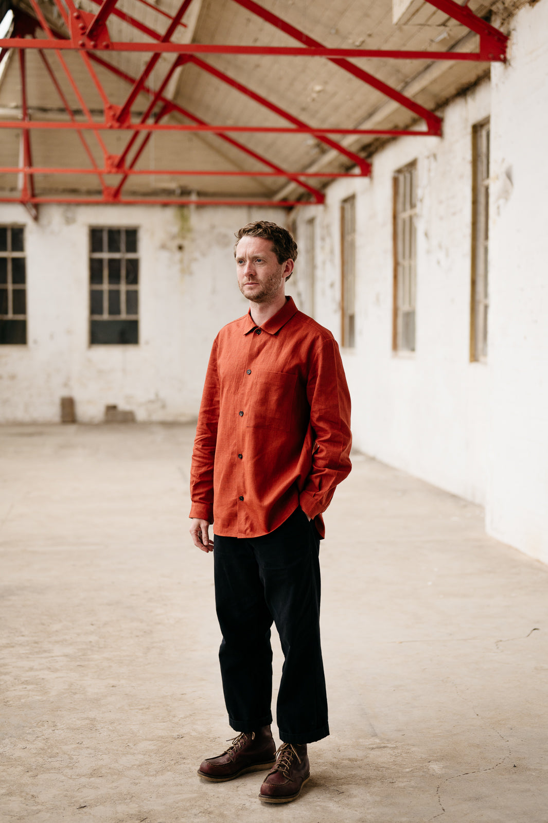 Man wearing an orange shirt and black pants standing in a large, empty warehouse with red metal beams.