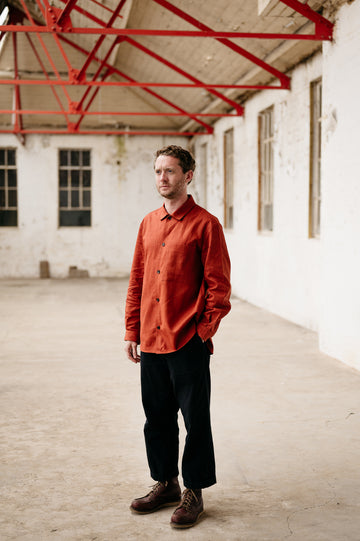 Man wearing an orange shirt and black pants standing in a large, empty warehouse with red metal beams.
