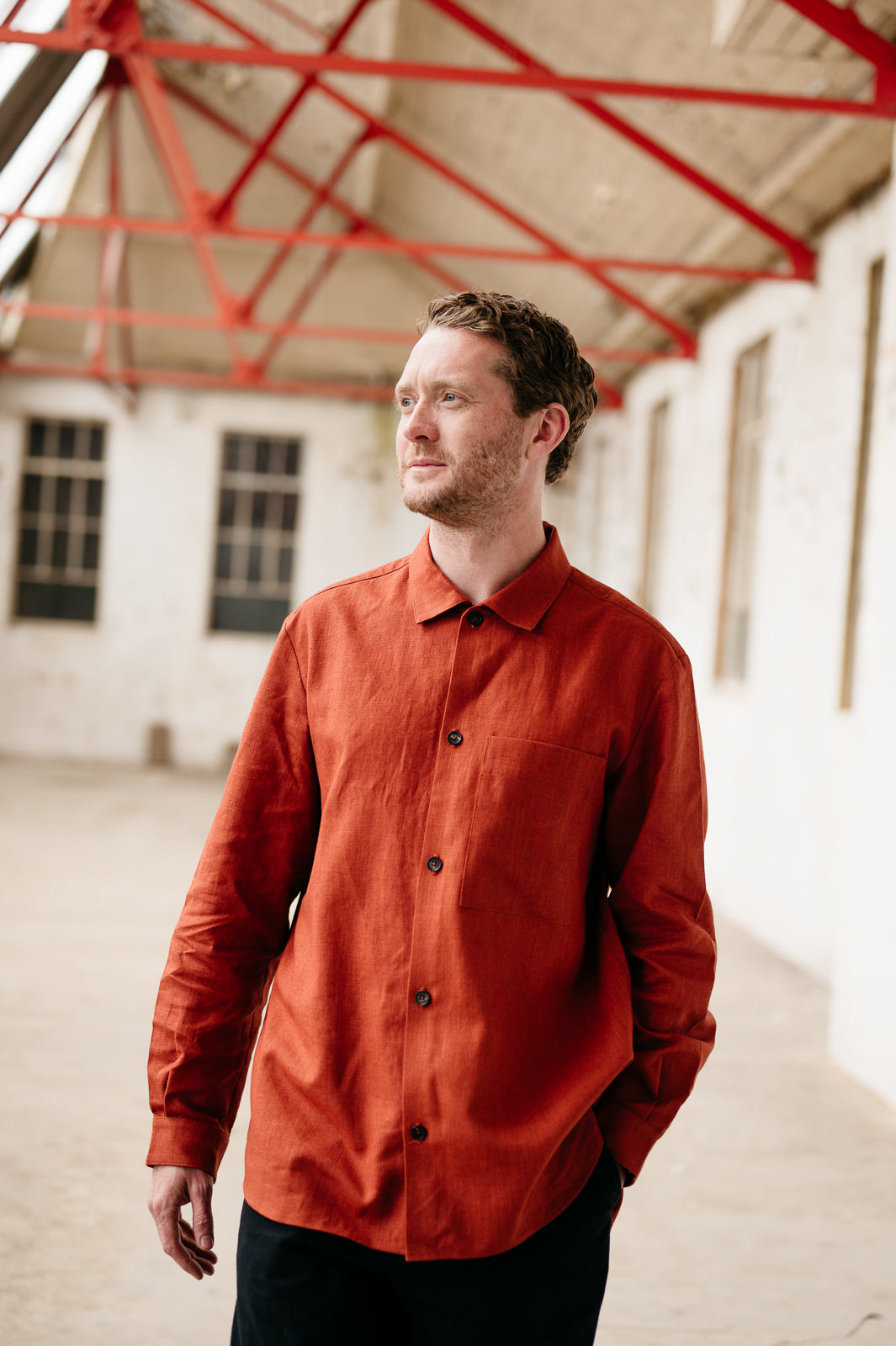 Man wearing a rust coloured shirt standing in an industrial setting with red structural elements.