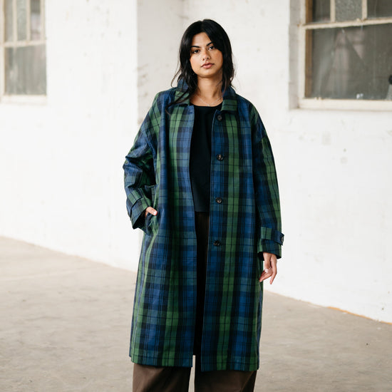 A woman stands in a white-walled industrial building wearing a long green and blue plaid coat over a black top and brown wide-leg pants, with red sneakers.