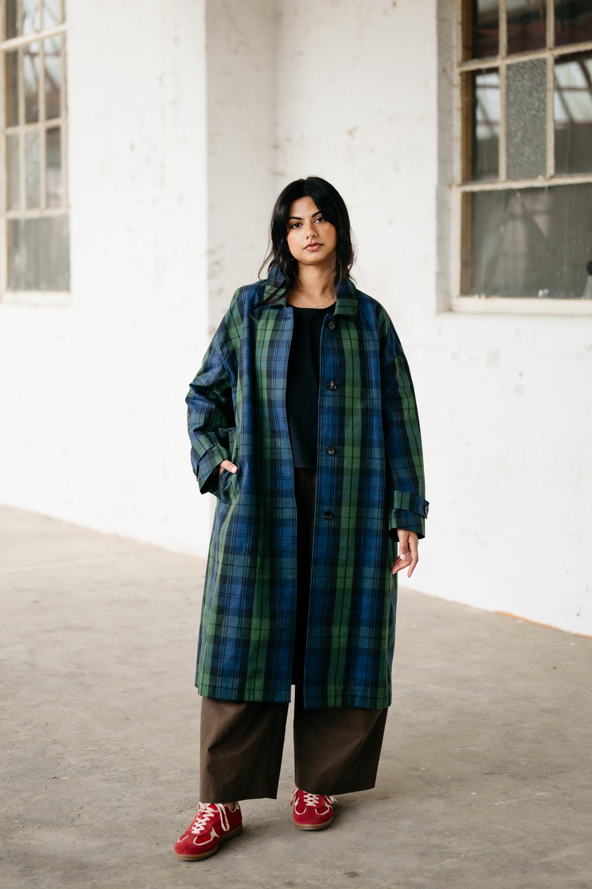 A woman stands in a white-walled industrial building wearing a long green and blue plaid coat over a black top and brown wide-leg pants, with red sneakers.