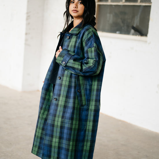 A woman wearing a long blue and green plaid coat, brown pants, and red shoes, standing in a bright, minimalist indoor space with a concrete floor and white walls.