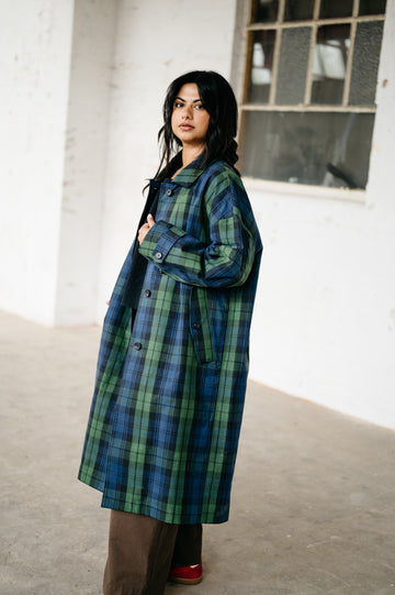 A woman wearing a long blue and green plaid coat, brown pants, and red shoes, standing in a bright, minimalist indoor space with a concrete floor and white walls.