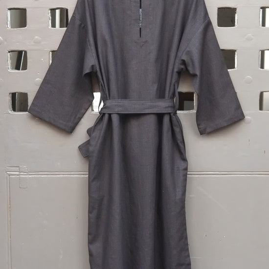 A black linen dress with long sleeves and a matching waist tie belt, hanging on a wooden hanger against a grey door with a square grid pattern.