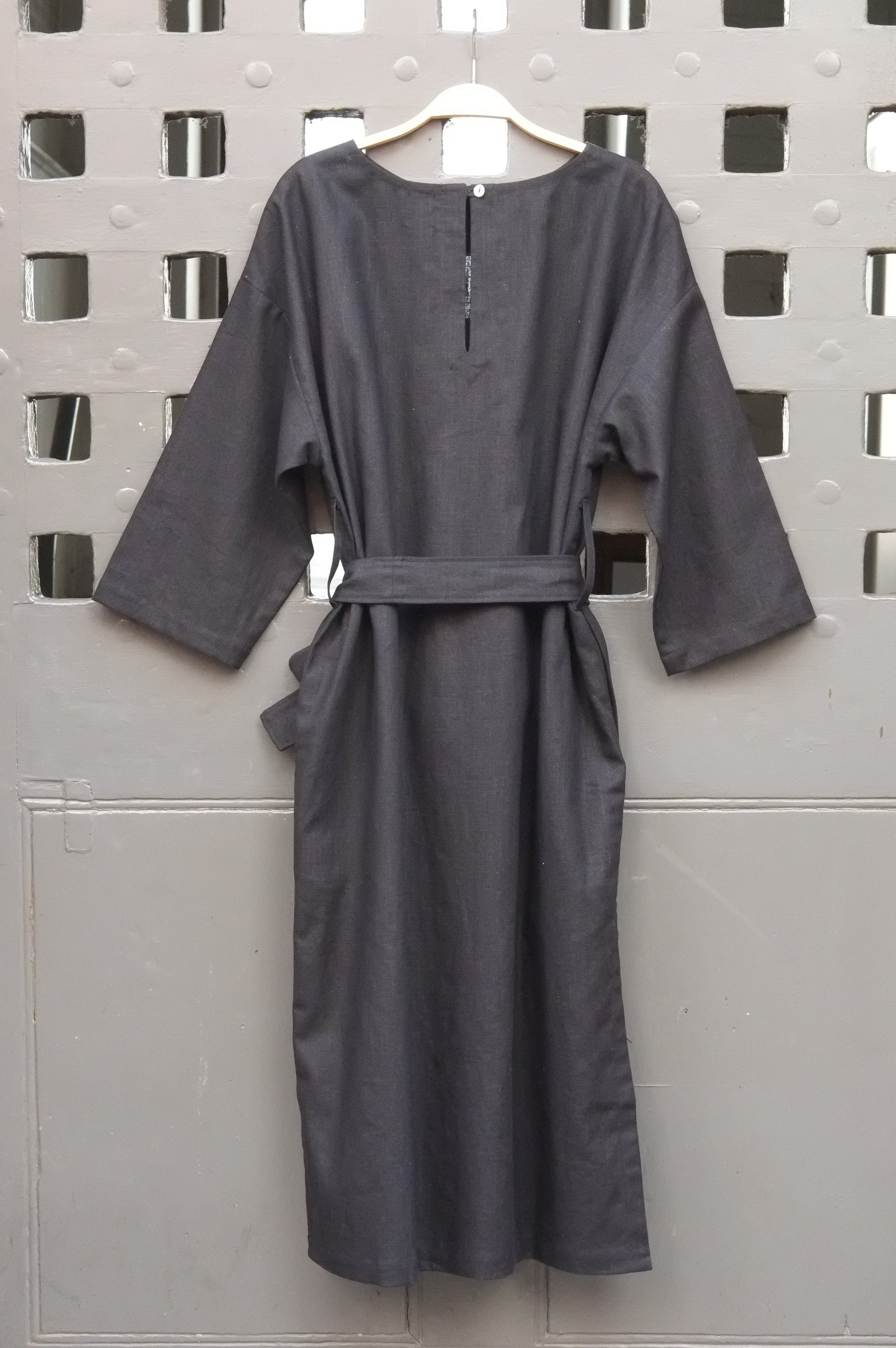 A black linen dress with long sleeves and a matching waist tie belt, hanging on a wooden hanger against a grey door with a square grid pattern.