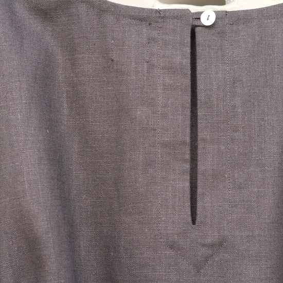 A close-up of a  linen dress hanging on a wooden hanger, featuring a split neckline with a central vertical slit and a single white button.