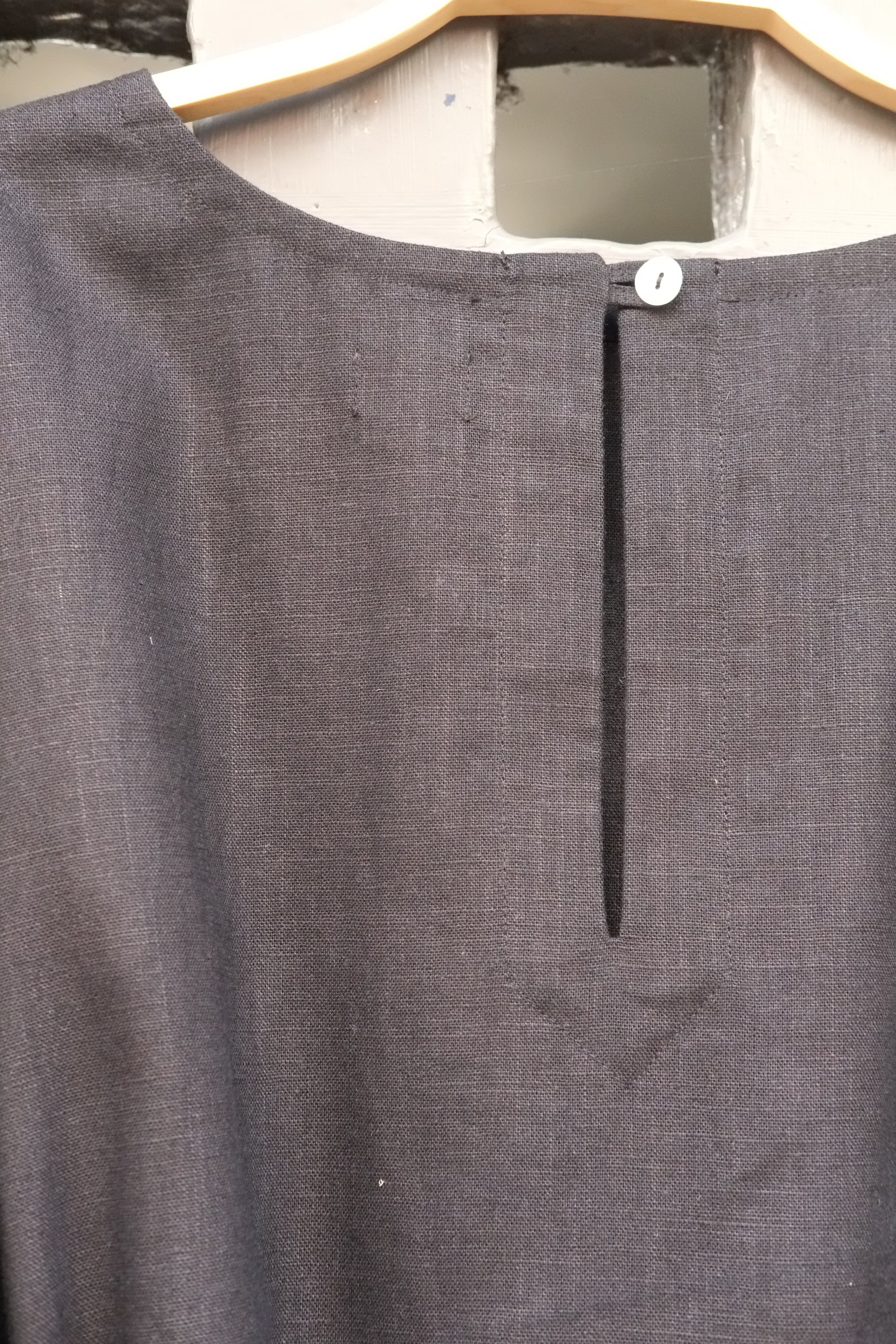 A close-up of a  linen dress hanging on a wooden hanger, featuring a split neckline with a central vertical slit and a single white button.