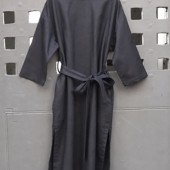 A black midi dress with three-quarter sleeves and a self-tie belt, hanging on a wooden hanger against a gray metal lattice screen.