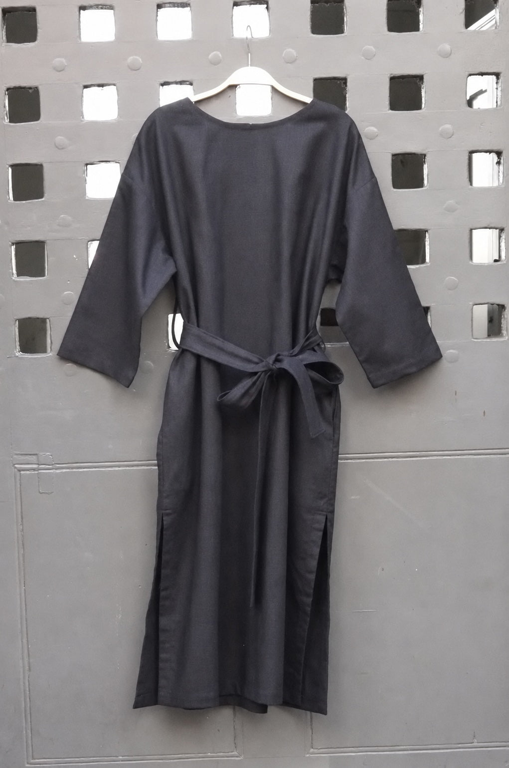 A black midi dress with three-quarter sleeves and a self-tie belt, hanging on a wooden hanger against a gray metal lattice screen.
