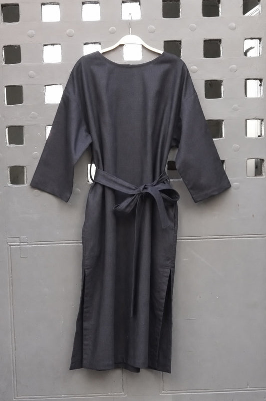 A black midi dress with three-quarter sleeves and a self-tie belt, hanging on a wooden hanger against a gray metal lattice screen.