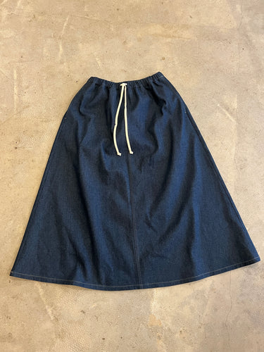 A-line indigo denim full length skirt with side pockets and a cream cord waist tie on a grey concrete background