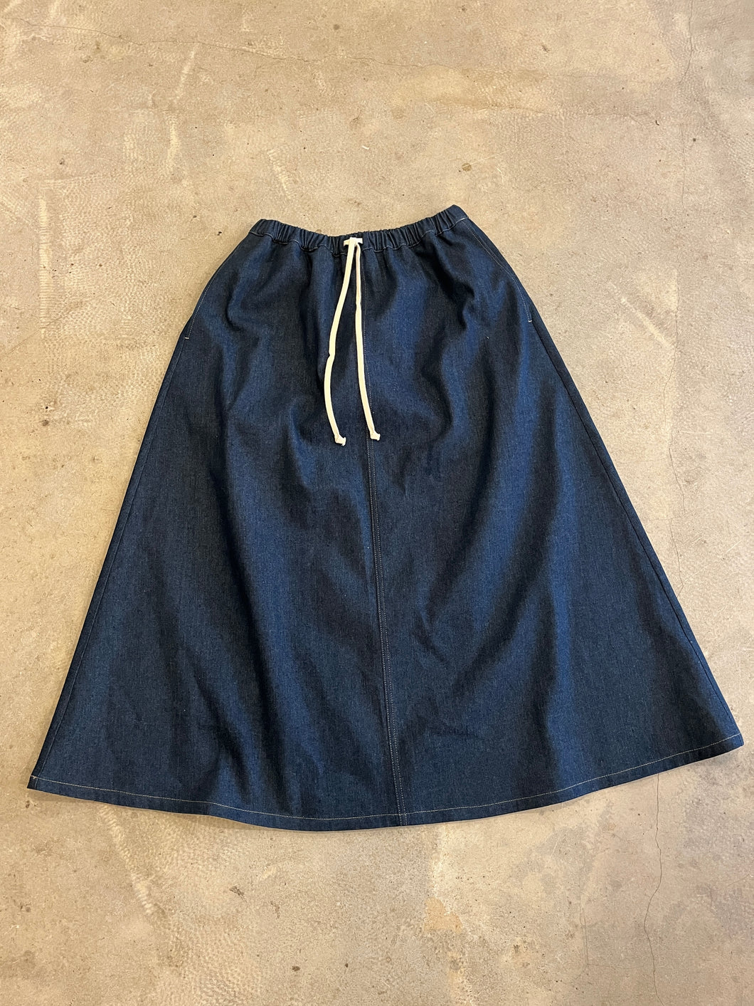 A-line indigo denim full length skirt with side pockets and a cream cord waist tie on a grey concrete background