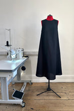 Load image into Gallery viewer, A black sleeveless, high neck dress with a deep v-back on a mannequin in a sewing studio setting, indicating a fitted garment made from ramie linen.
