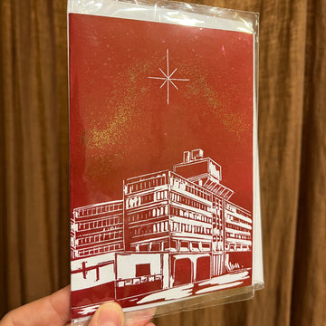 crimson red Christmas card with gold glitter featuring the UEA in Norwich and a star in the sky, held by a hand against a brown curtain