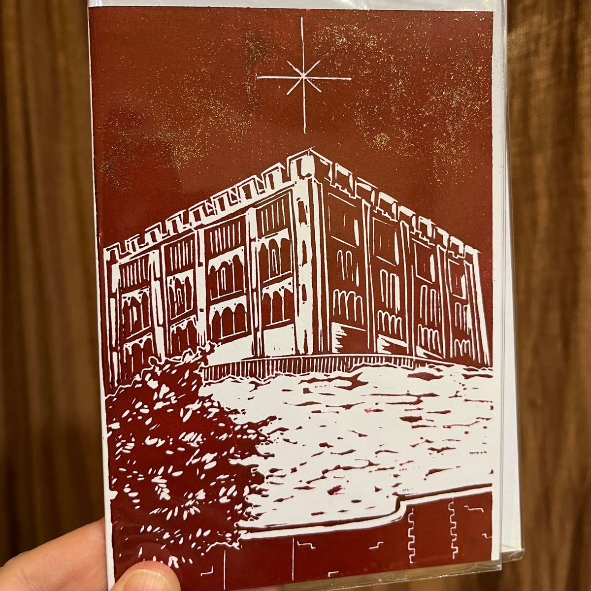 A hand holding an art card featuring a white architectural illustration of a Norwich Castle against a dark reddish-brown background, still sealed in clear plastic packaging, with a wooden surface behind it.