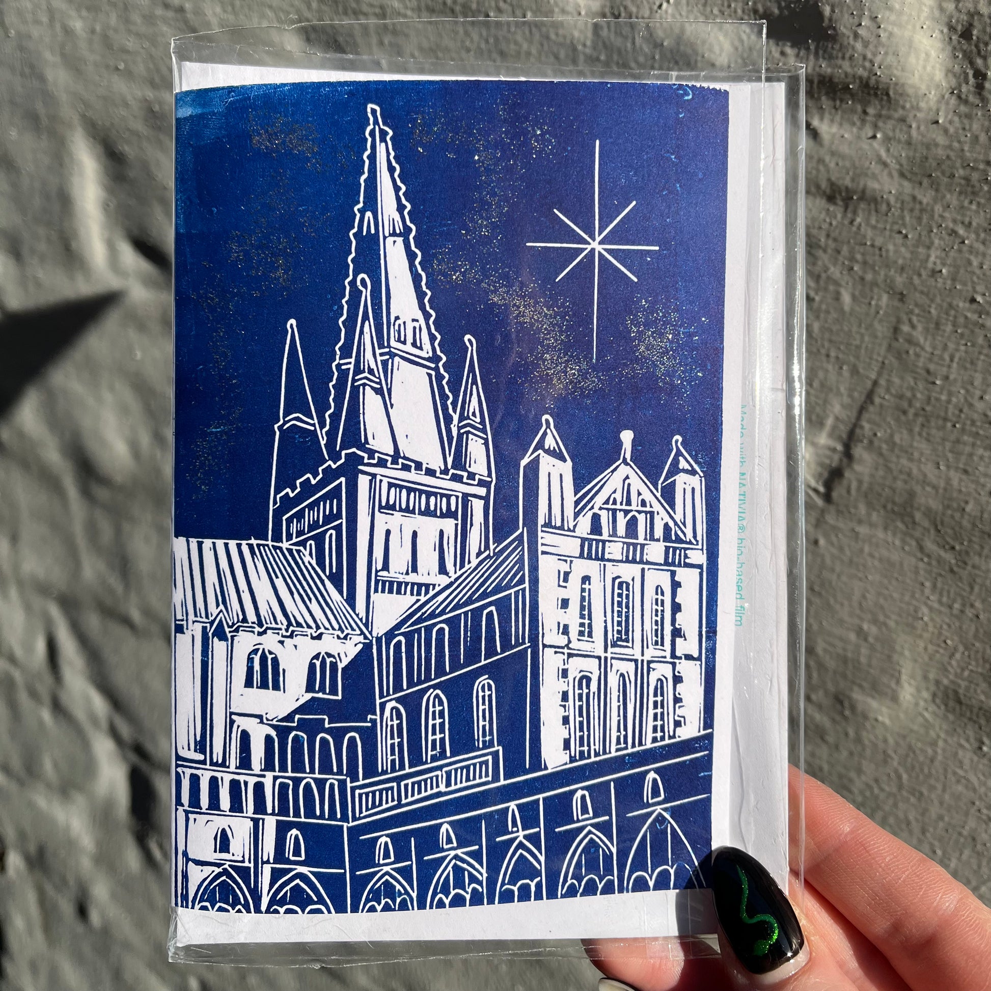 Card with a blue and white illustration of a cathedral held by a hand against a stone wall.