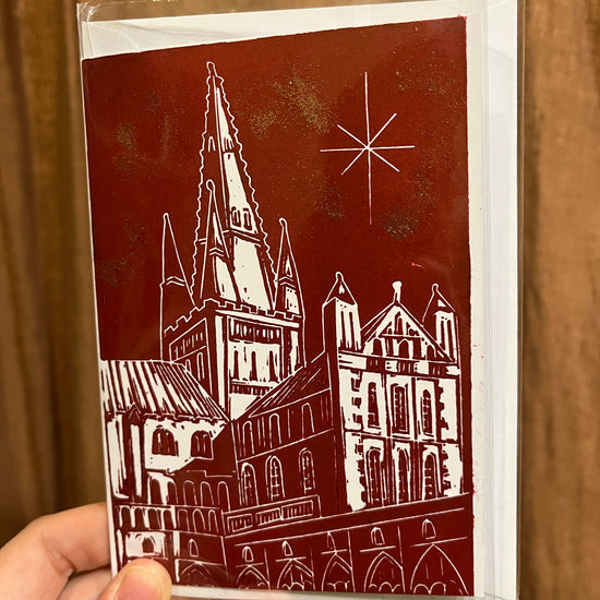 crimson Christmas card with a white Lino printed illustration of Norwich Cathedral with a star in the sky, held by a hand infant of a brown curtain