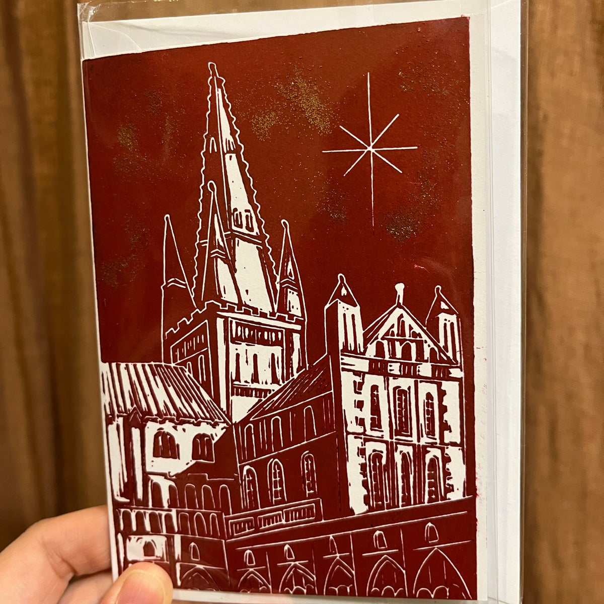 crimson Christmas card with a white Lino printed illustration of Norwich Cathedral with a star in the sky, held by a hand infant of a brown curtain