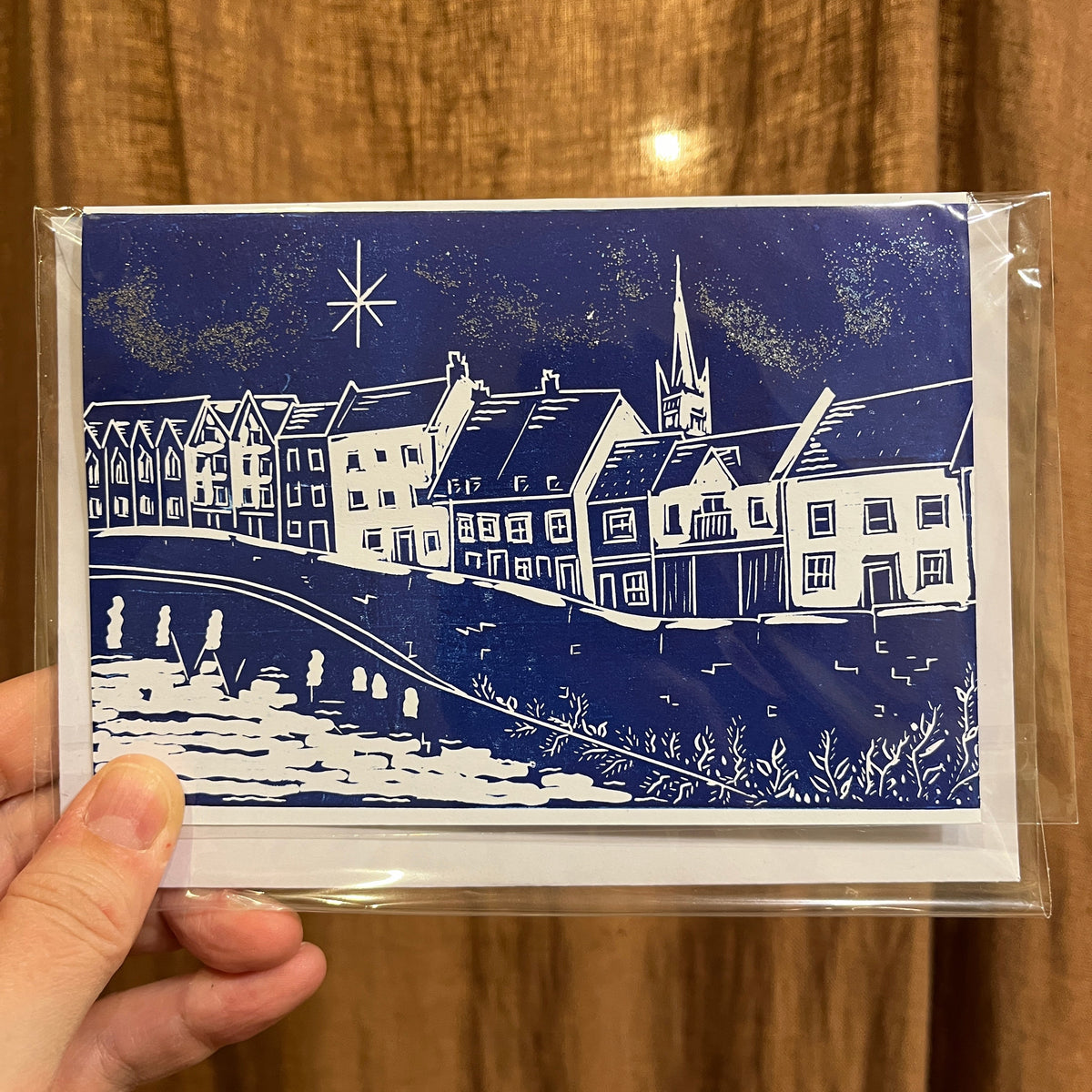 A hand holding a clear plastic sleeve containing a square art print or card featuring a blue and white block-print style illustration of river houses, a church spire, and a starry sky against a brown wood-textured background.