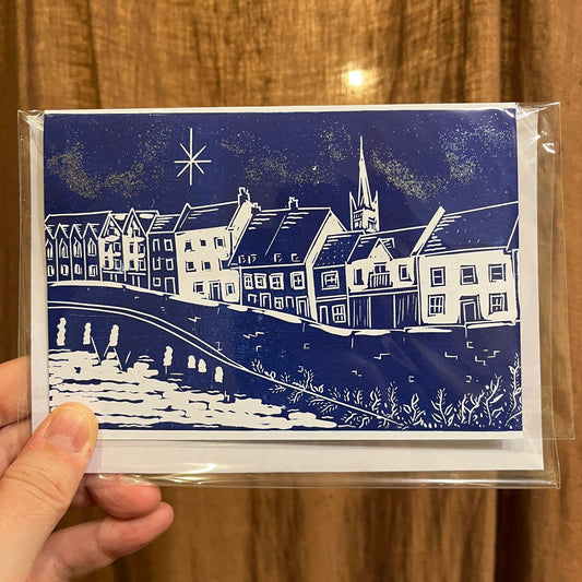 A hand holding a clear plastic sleeve containing a square art print or card featuring a blue and white block-print style illustration of river houses, a church spire, and a starry sky against a brown wood-textured background.