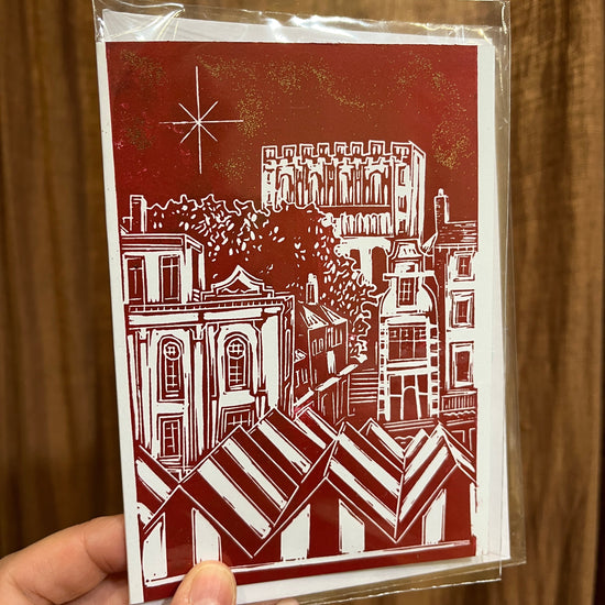 crimson red Christmas card featuring a view from the top of Norwich Market in white ink from a lino print, held by a hand infant of a brown curtain