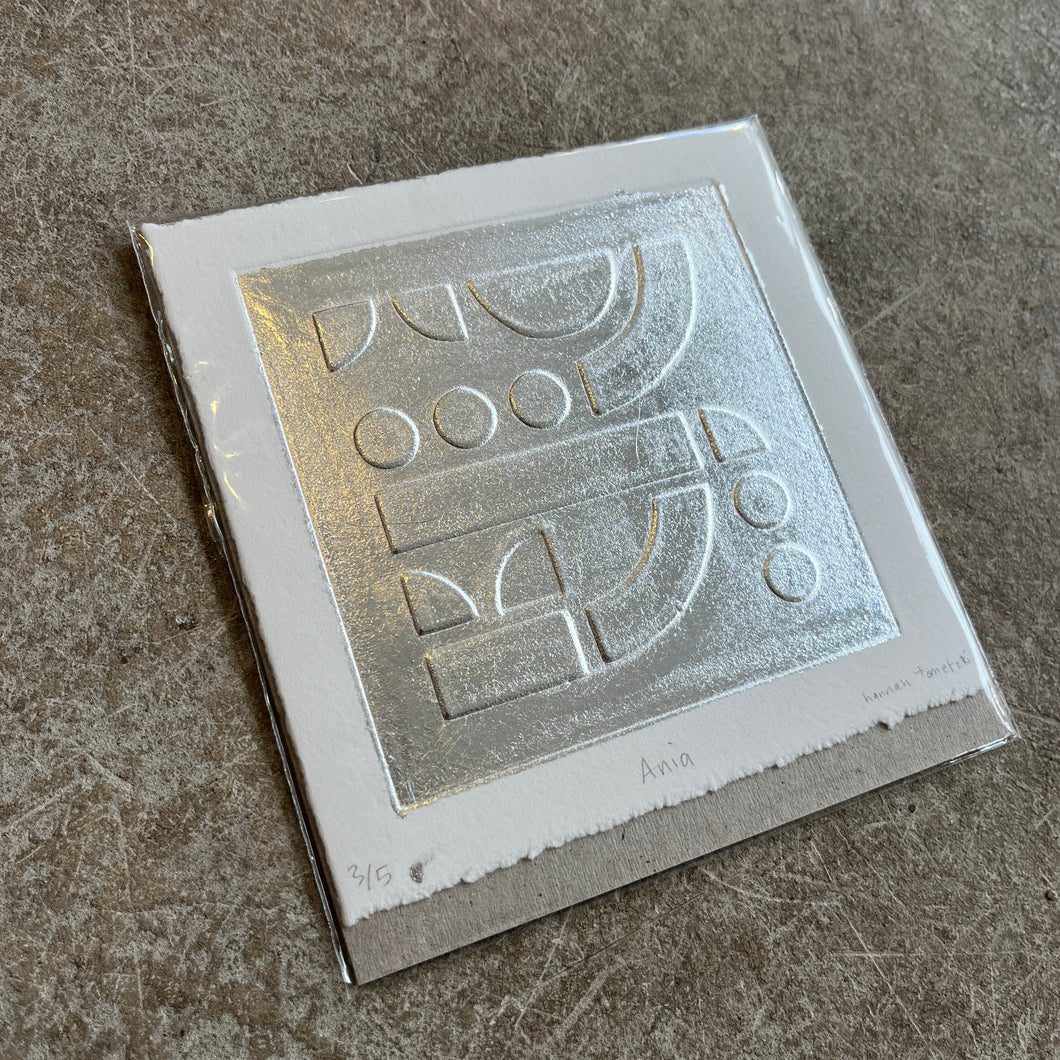 Silver leaf embossed geometric abstract print with a white boarder on heritage cotton paper. Square shaped on a grey concrete background