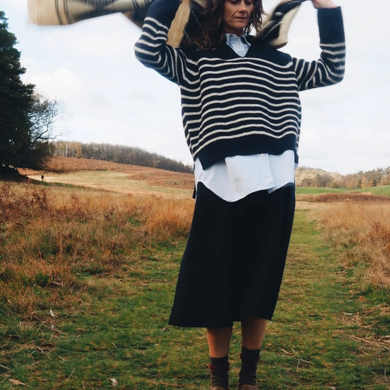 Model wears navy and cream stripe v neck jumper with blue shirt underneath and navy blue skirt while waving a scarf behind her in a country field setting