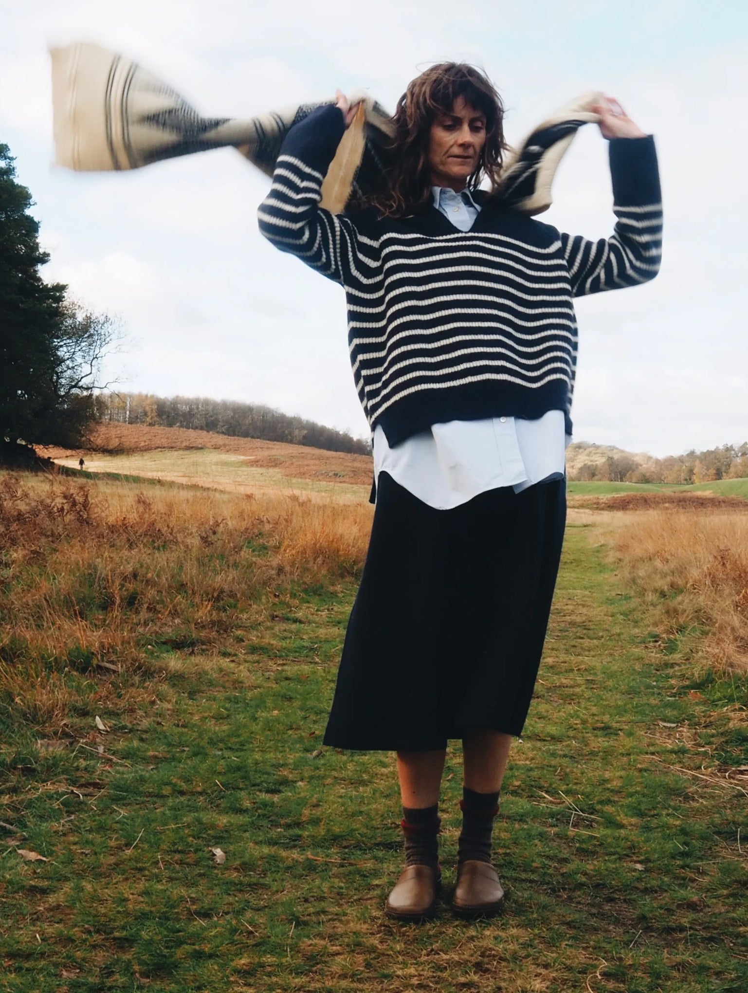 Model wears navy and cream stripe v neck jumper with blue shirt underneath and navy blue skirt while waving a scarf behind her in a country field setting