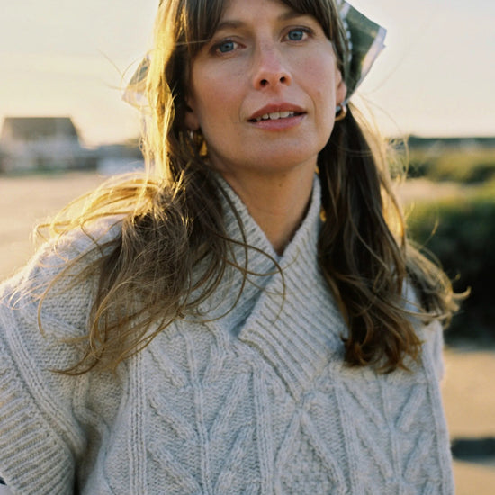 Woman wearing a cable knit v neck pale cream sweater with a blurred outdoor background