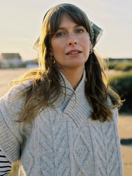 Woman wearing a cable knit v neck pale cream sweater with a blurred outdoor background