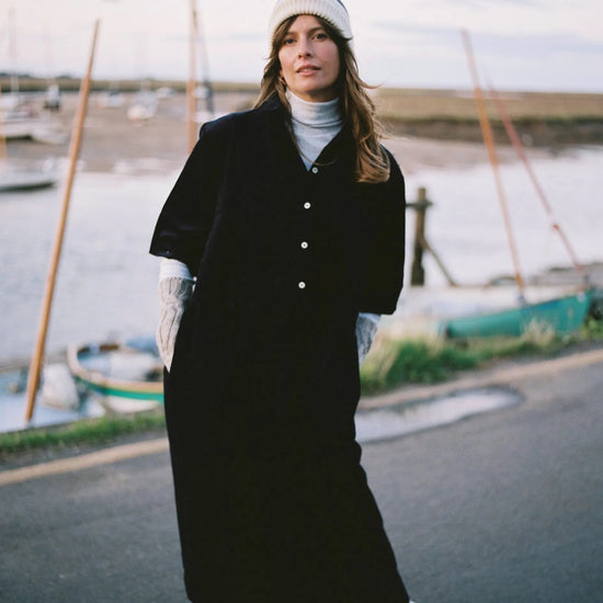 Person in a long navy cord dress with arm arm warmers on and knit hat stretching outdoors by a body of water with boats