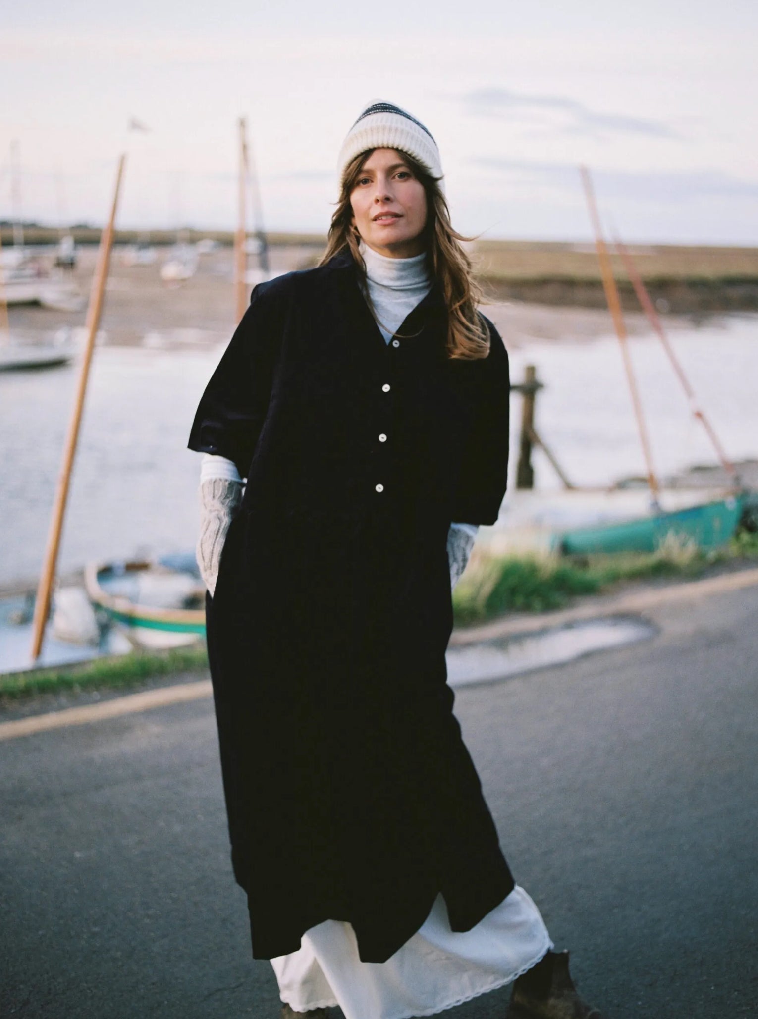 Person in a long navy cord dress with arm arm warmers on and knit hat stretching outdoors by a body of water with boats