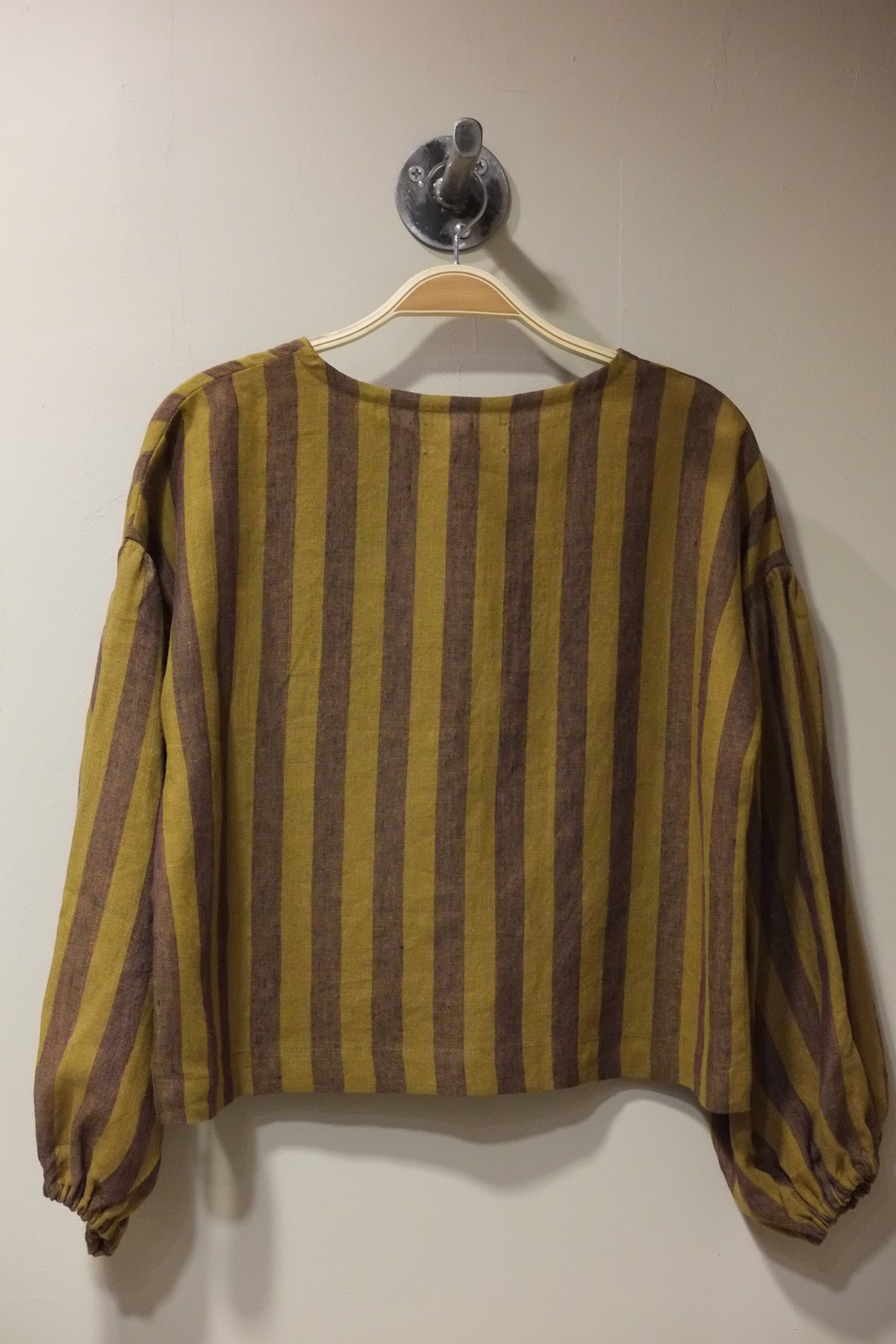 A long-sleeve striped top in mustard yellow and brown hanging on a wooden hanger against a plain light wall.